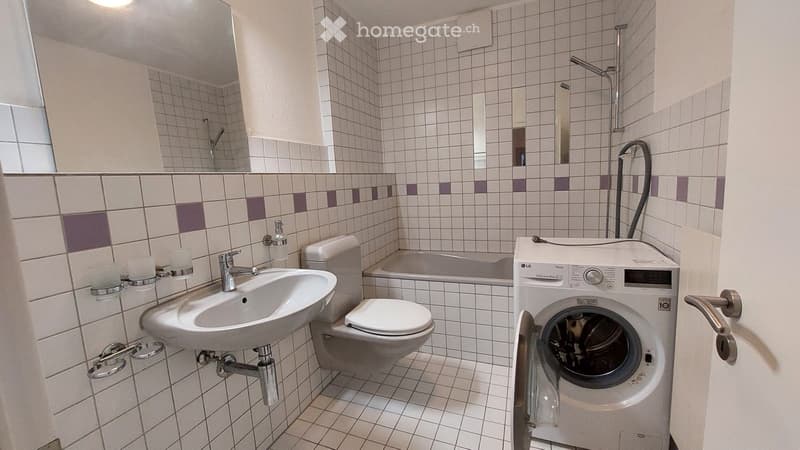 Bathroom with white tiles, a sink, toilet, bathtub, and a washing machine. Features a shower area with a glass enclosure and a mirror above the sink.
