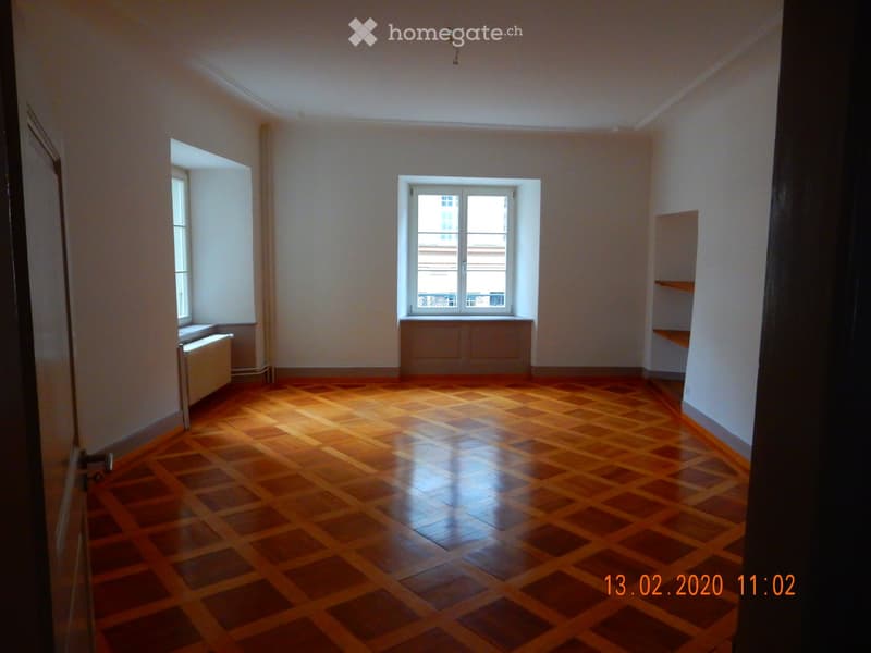 Empty room with wooden checkered flooring, white walls, and two windows allowing natural light. The room features built-in shelving on one wall and a radiator under one window.