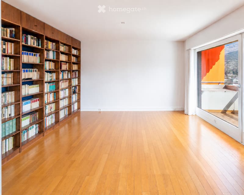 A spacious room with wooden flooring and a large bookshelf filled with books, featuring large windows with a scenic mountain view.