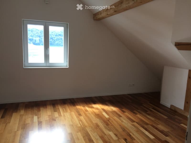 Empty attic room with wooden flooring, exposed wooden beams, and a large window offering a view of greenery.