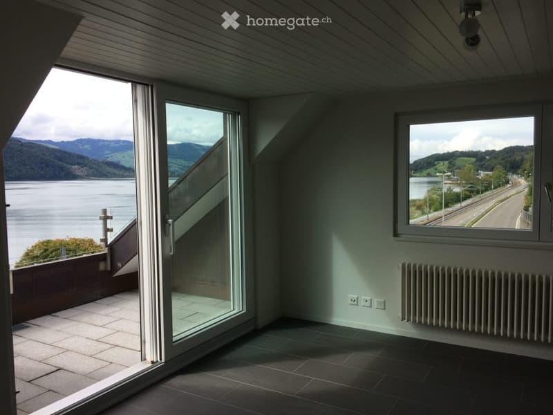 Empty room with large windows and a sliding glass door leading to a balcony, offering a scenic view of a lake and mountains.