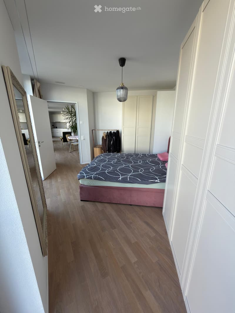 Spacious bedroom with wooden flooring, a large bed with a patterned duvet, a wardrobe, a coat rack, and an open doorway leading to a kitchen area.