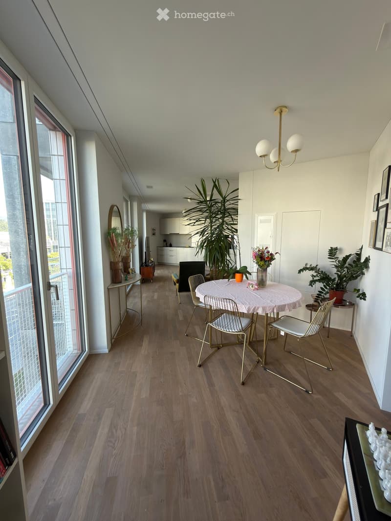 Open-plan living and dining area with wooden flooring, large windows, a round dining table with chairs, a side table with a mirror, and several potted plants.