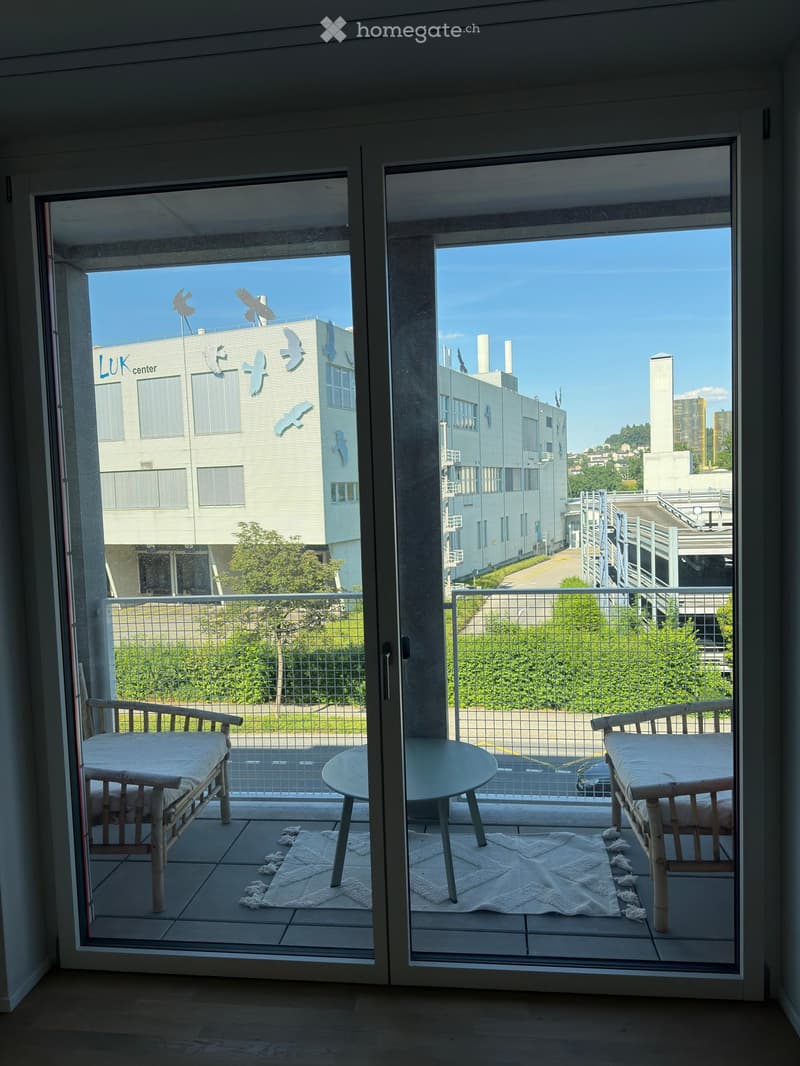 Balcony with two chairs, a small round table, and a white rug. The view shows a modern building with bird sculptures and a parking area.