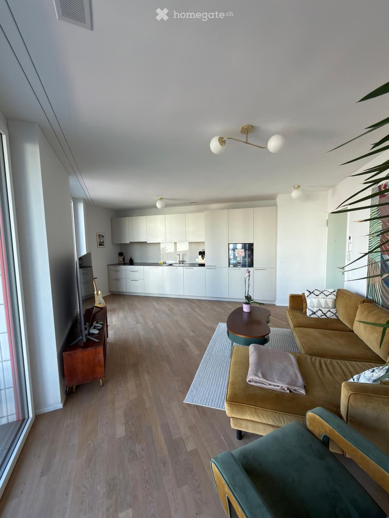 Modern living room with a beige sofa, coffee table, small round table, and a guitar on a stand. The room features light wooden flooring, white kitchen cabinets, and a sleek refrigerator. A television is mounted on the wall, and a floor lamp provides ambie