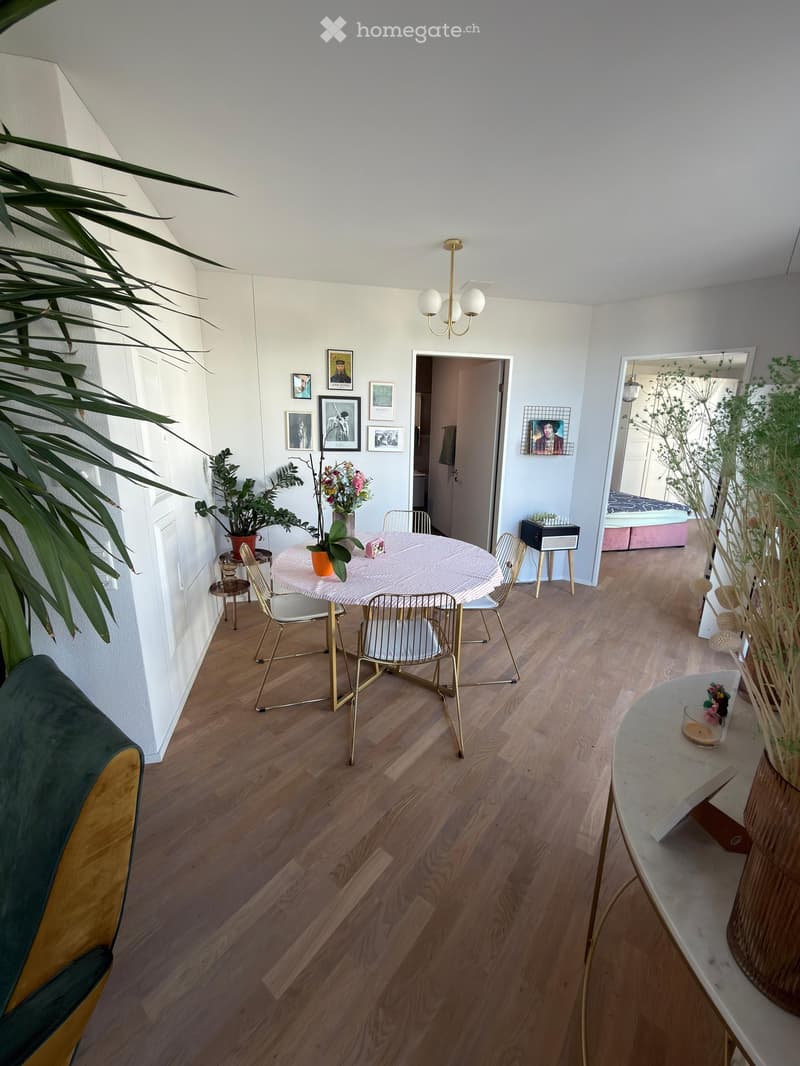A modern, well-lit living space with wooden flooring, a round dining table with chairs, a green and yellow sofa, and several potted plants. The room features artwork on the walls and an open doorway leading to another room.