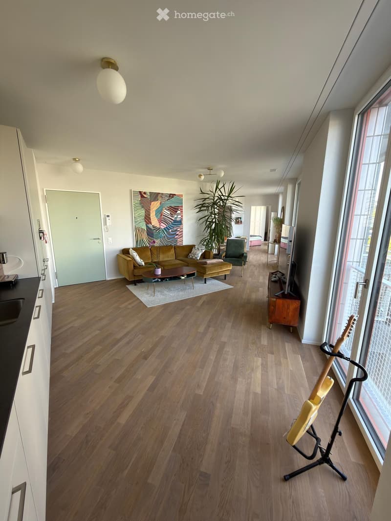 Open-plan living and dining area with wooden flooring, a mustard-colored sofa, coffee table, ottoman, armchair, guitar on a stand, and a modern kitchen area with a sink and cabinet.