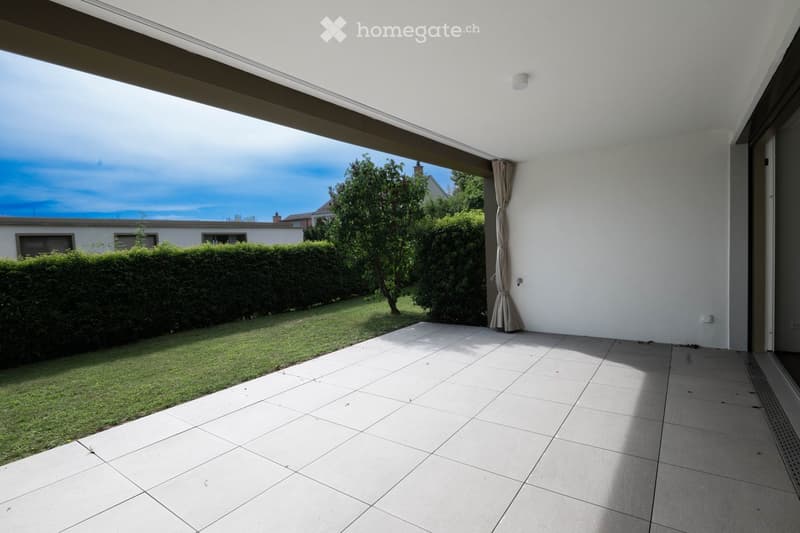 Spacious balcony with tiled flooring, white walls, and a view of a well-maintained garden with hedges and trees.