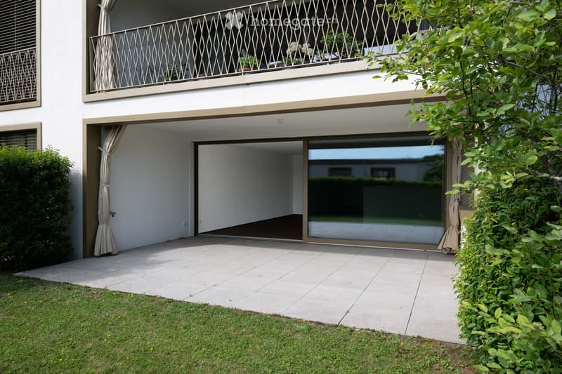 Modern outdoor area with tiled patio, sliding glass doors leading to a white building with balconies, greenery, and neatly trimmed hedges.