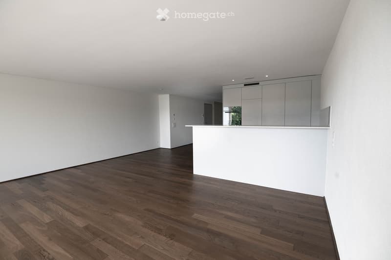 Empty room with white walls, dark wooden flooring, and a white counter with cabinets in the background.