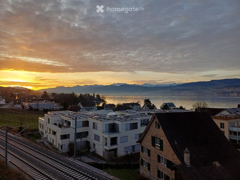A panoramic view of a town with modern and traditional houses, a train track, and a lake with mountains in the background during sunset.
