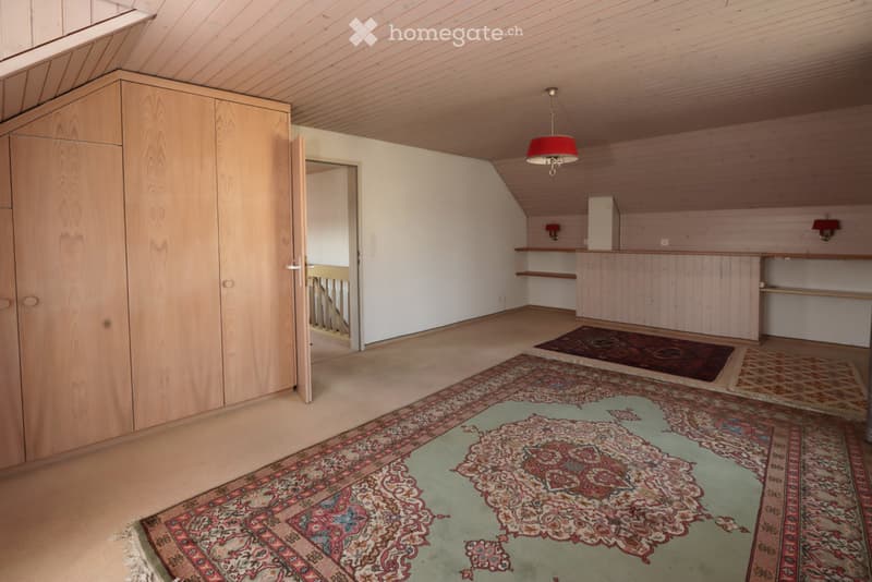 An unfurnished attic room with wooden walls and ceiling, featuring built-in wooden shelving units, a hanging red lamp, and several patterned rugs on the floor.