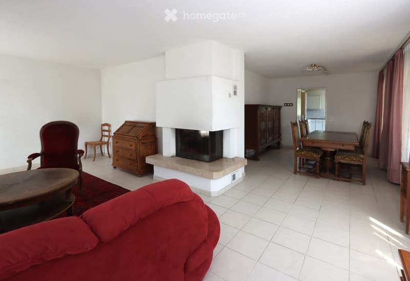 A spacious living room with a red sofa, coffee table, fireplace, dining area with a table and chairs, and a wooden desk.