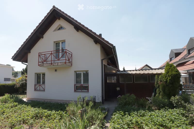 Single-story house with white walls, red-framed windows, and a balcony on the upper level. The house features a well-maintained garden with various plants and shrubs in the foreground.
