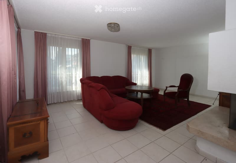 Living room with red sofa, wooden coffee table, red armchair, and wooden cabinet. The room features tiled flooring, large windows with curtains, and a chandelier.