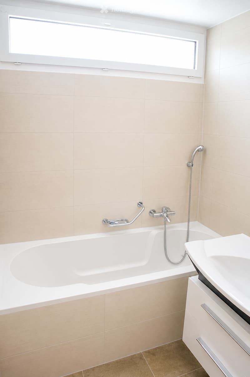 Modern bathroom with a bathtub, faucet, sink, and cabinet. Light beige tiles and a large window provide natural light.