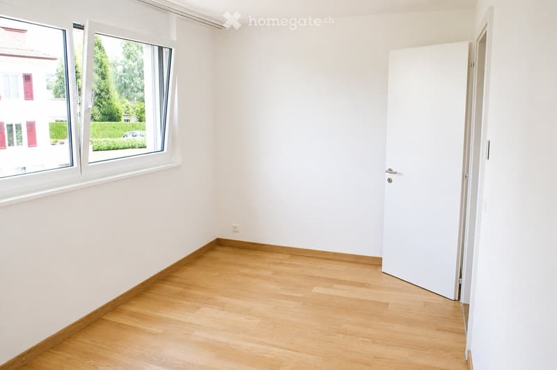 Empty room with light wooden flooring, white walls, and large windows offering a view of greenery and neighboring buildings.