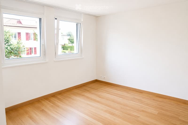 Empty room with light-colored walls and wooden flooring, featuring two large windows with white frames and blinds.
