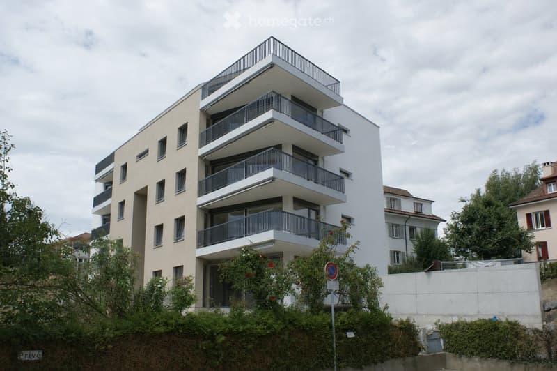 Modern multi-story residential building with balconies and large windows, surrounded by greenery and neighboring structures.