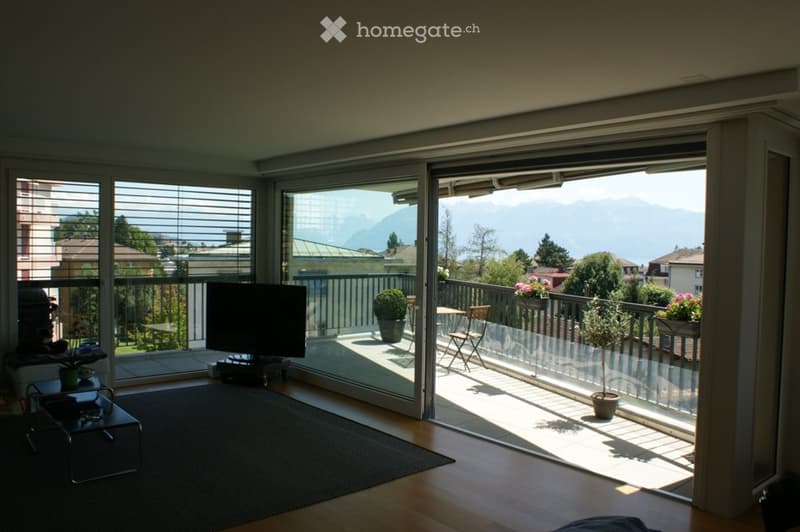 Bright living room with large sliding glass doors opening to a balcony, featuring a sofa, coffee table, television, and potted plants. The balcony has outdoor furniture and offers a scenic mountain view.