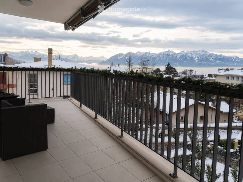 Balcony with tiled floor, black metal railing, and a view of snow-capped mountains and a town below.