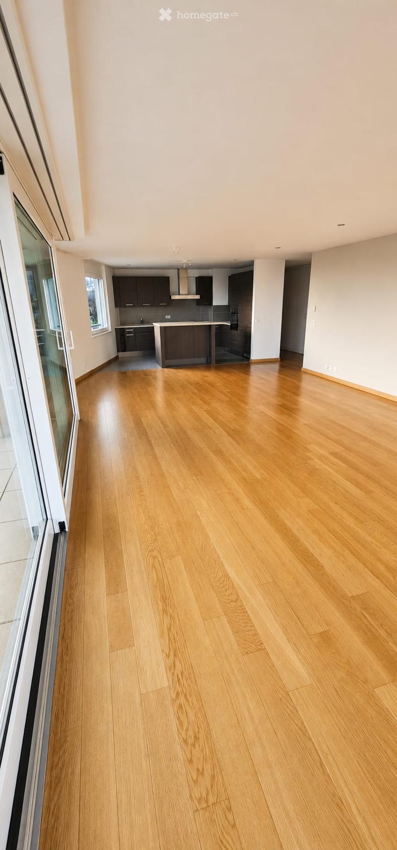 Empty room with light wooden flooring, white walls, and large windows. The adjacent kitchen area is visible in the background with cabinets and a countertop.