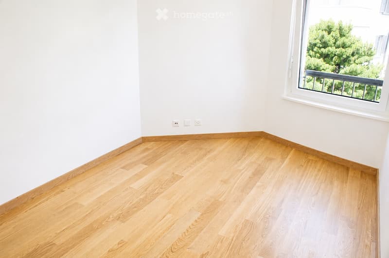 Empty room with light wooden flooring and white walls, featuring a large window with a view of greenery and a balcony railing visible outside.