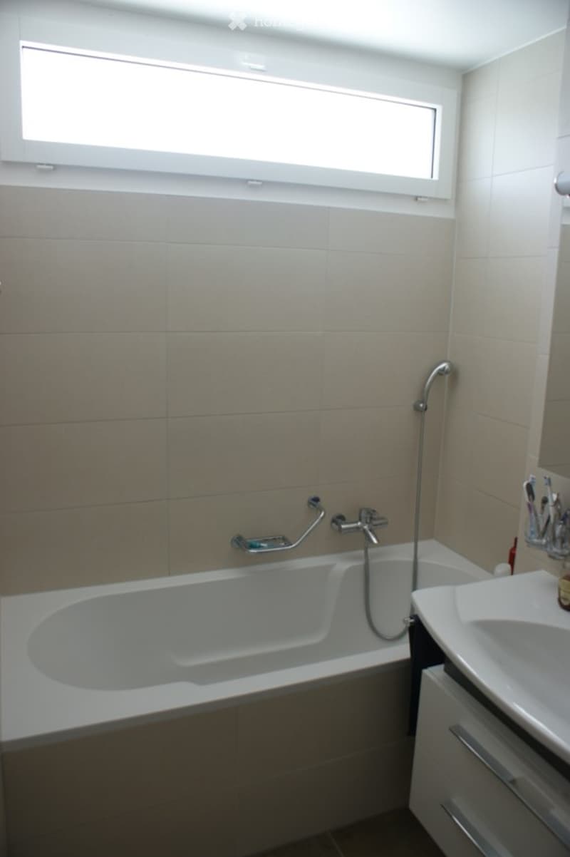 Modern bathroom with a bathtub, sink, and a large window providing natural light. The walls are tiled in a neutral color, and the fixtures are sleek and contemporary.