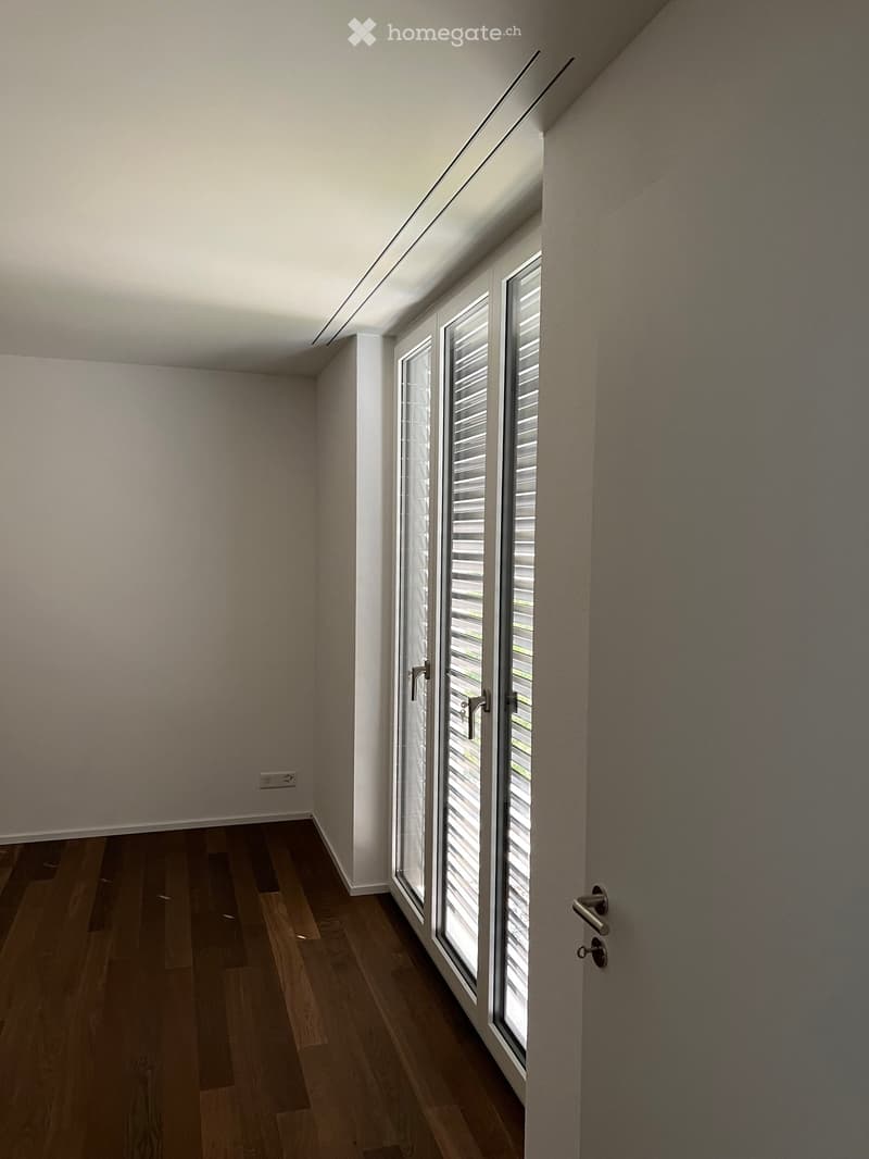 Empty room with wooden flooring, white walls, large window with blinds, and a door with a handle.