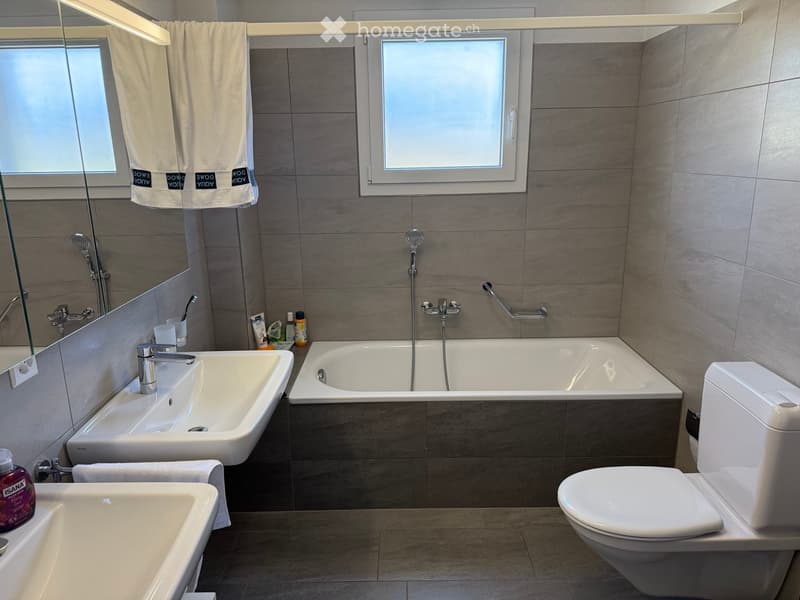Modern bathroom with two sinks, a bathtub, a toilet, a large mirror, and a towel hanging on a rack. The walls are tiled in a neutral color, and the floor is made of light-colored tiles.