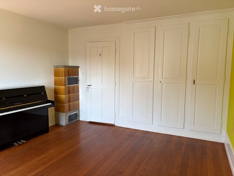 Empty room with wooden flooring, white walls, and a black piano against one wall. Three white doors are visible, and a small built-in cabinet with a vent is present.