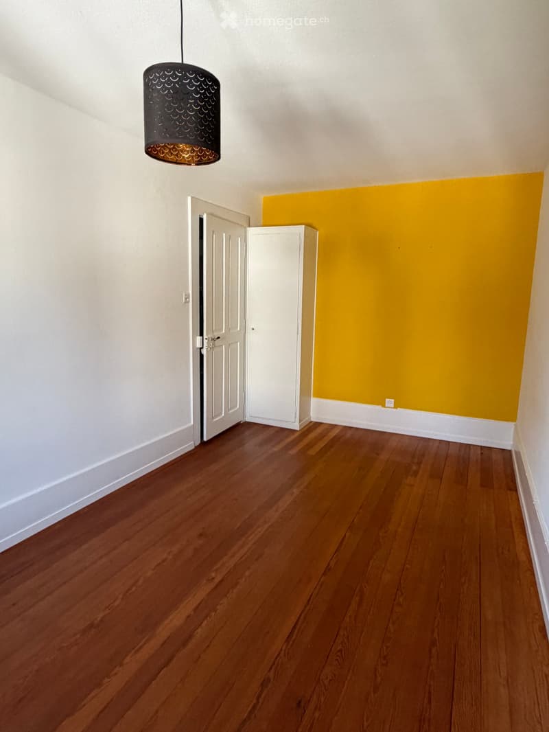 Empty room with wooden flooring, white walls, a hanging light fixture, a white cabinet, and a partially open door leading to another space.