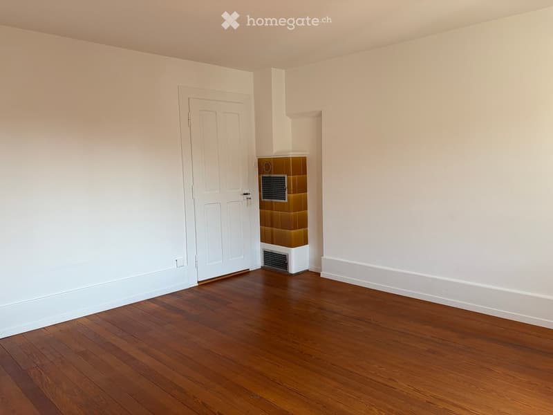 Empty room with wooden flooring, white walls, and a white door. A small tiled section is visible near the door.
