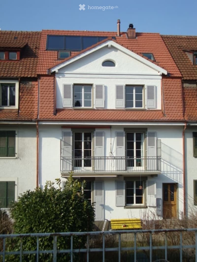 Two-story house with a red-tiled roof, white exterior, multiple windows with shutters, and a small balcony on the first floor.