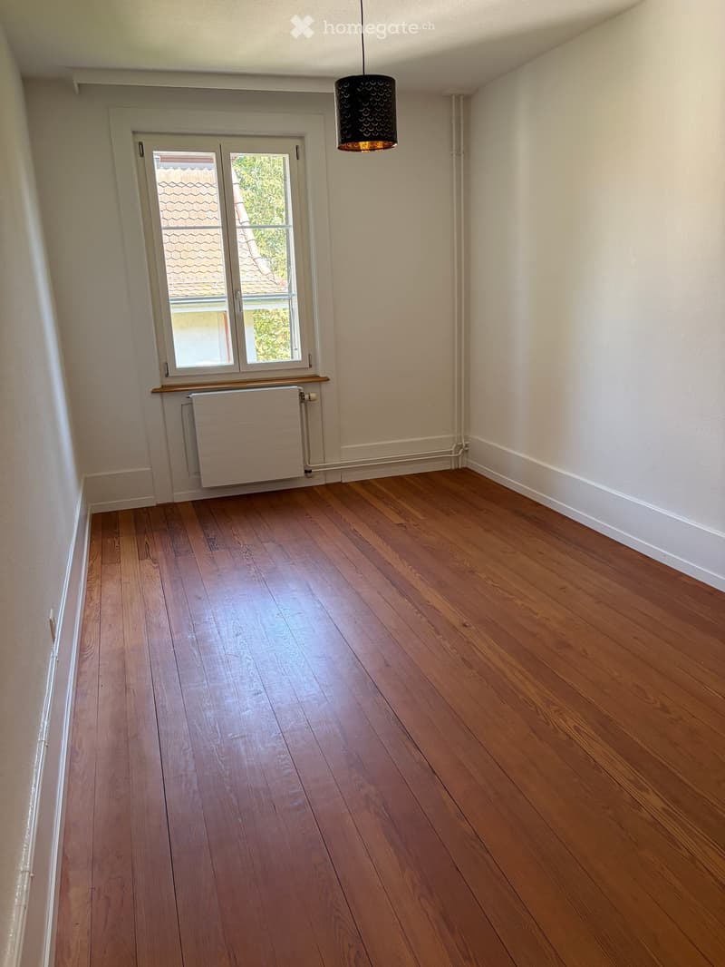 Empty room with wooden flooring, white walls, a window with a radiator below, and a hanging light fixture.