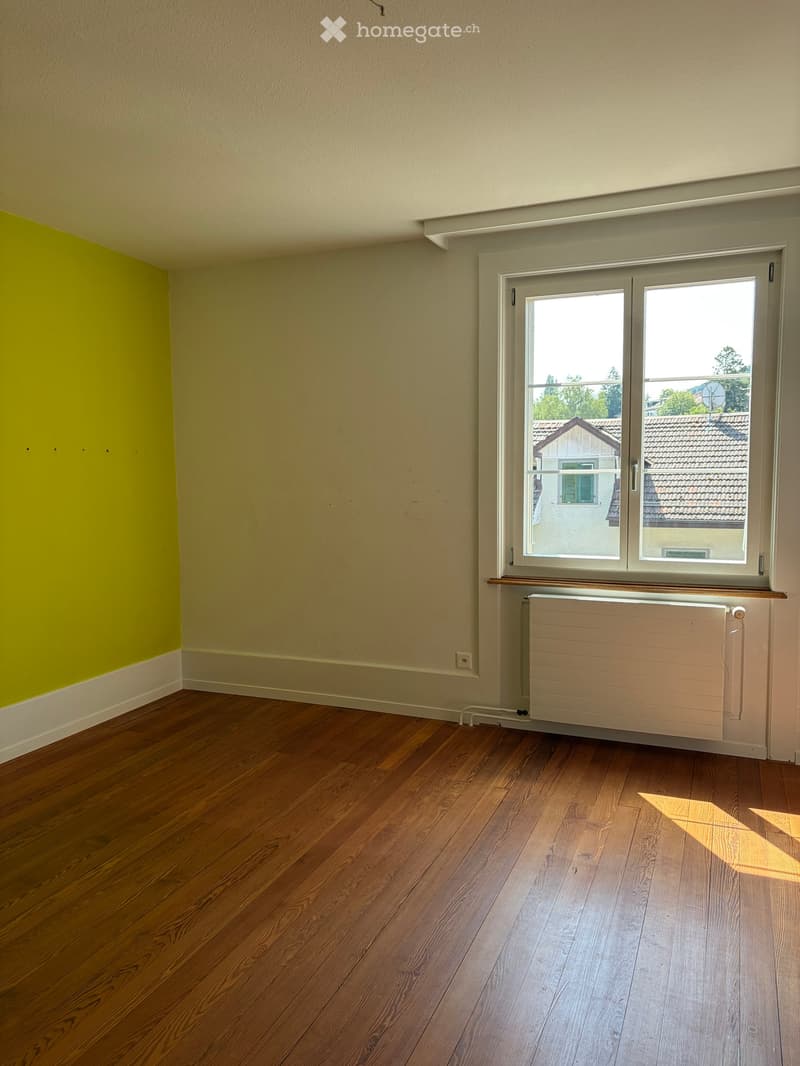 Empty room with wooden flooring, light-colored walls, and a large window with a radiator below it.