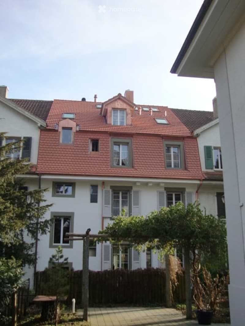 Two-story residential house with red-tiled roof, white walls, multiple windows, and a small garden area with a wooden pergola and seating.