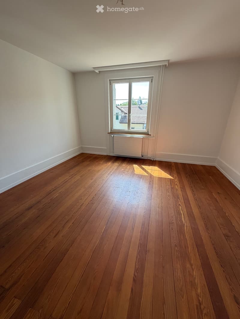 Empty room with wooden flooring, white walls, and a window with a radiator below it.