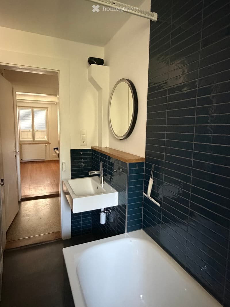 Modern bathroom with dark blue tiles, white bathtub, white sink, wooden shelf, and round mirror.