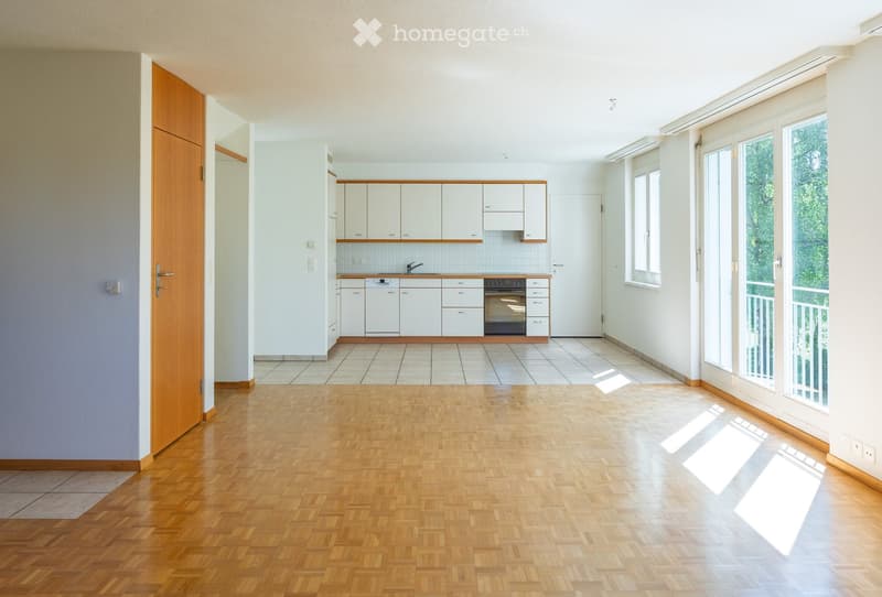 Bright, open-plan room with wooden flooring, white cabinets, and a small kitchen area featuring a sink, faucet, oven, and dishwasher. Large windows and a sliding glass door lead to a balcony.