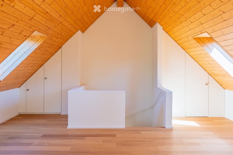 An empty attic space with wooden floors and slanted wooden ceilings, featuring built-in white cabinets and natural light from skylights.