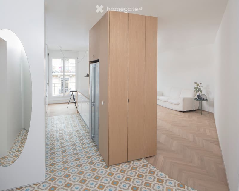 Modern, open-plan living space with a white sofa, a small round table, a large mirror, a refrigerator, and a wooden cabinet. The floor features patterned tiles in the hallway and wooden flooring in the living area. A balcony is visible through large windo