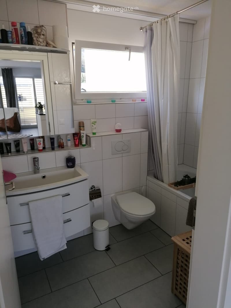 Modern bathroom with white tiles, a sink, mirror, toilet, bathtub, and various toiletries on the counter. Features a window with blinds and a small wooden shelf near the bathtub.