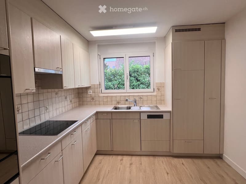 Modern kitchen with light wood cabinets, a sink, a dishwasher, and a stove, featuring a window with a view of greenery outside.