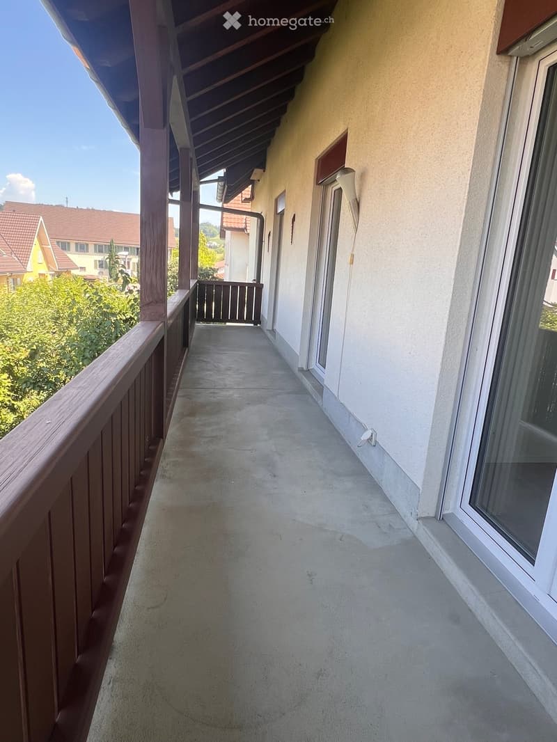 Balcony with wooden railing, concrete floor, and a view of greenery and neighboring houses.