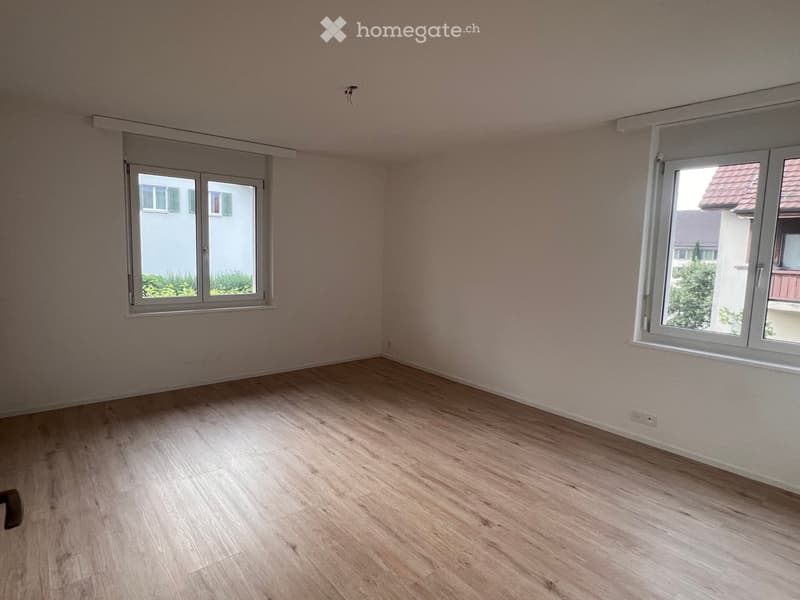 Empty room with light wooden flooring and white walls, featuring two windows with white frames and blinds.
