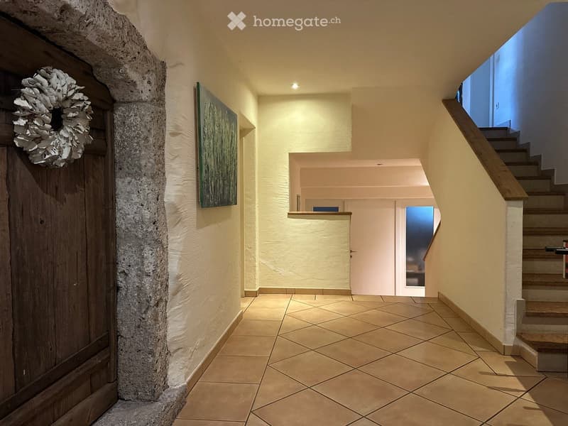 A well-lit hallway with tiled flooring, a stone pillar, a wooden door with a decorative wreath, and a staircase leading upwards.