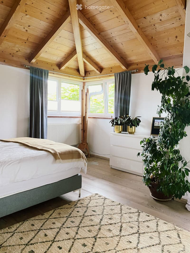 Cozy attic bedroom with wooden slanted ceilings, large windows, a bed with a green headboard, a nightstand with plants, and a large leafy plant in a pot.