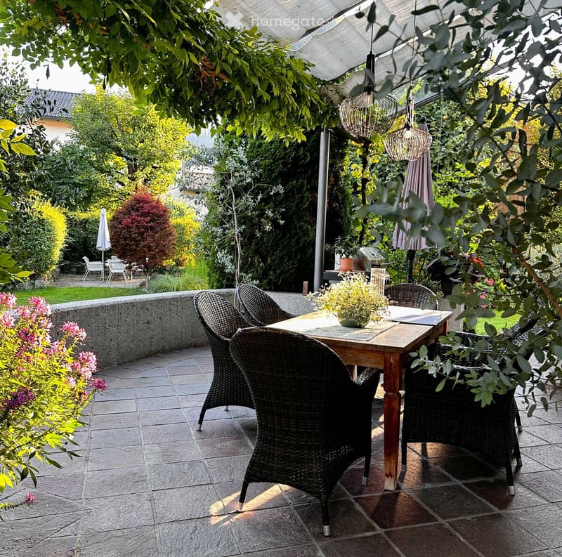 A shaded outdoor patio with a wooden table, wicker chairs, hanging lanterns, and potted plants, surrounded by greenery and a stone wall.