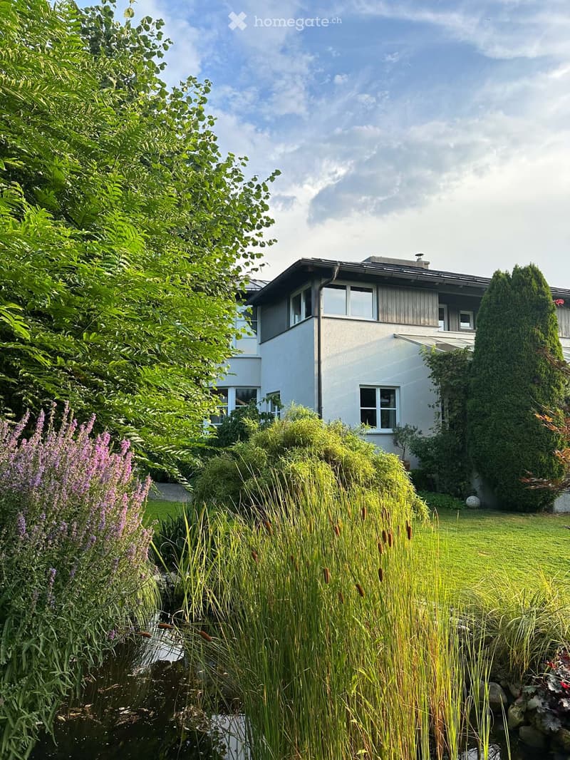 A modern two-story house with white walls and large windows, surrounded by a well-maintained garden with tall grasses, shrubs, and flowering plants.